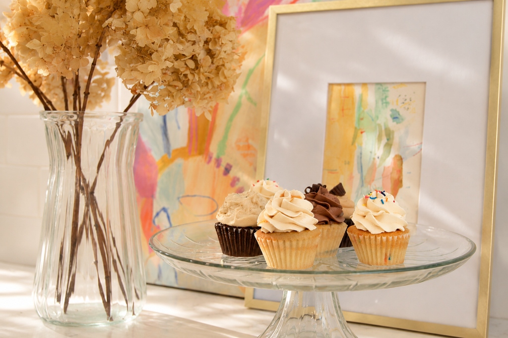 Gourmet Cupcakes on Glass Cake Stand next to Vase and Artwork